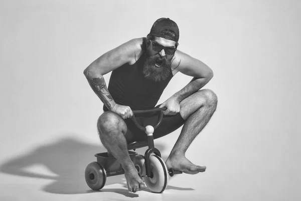 Бородатий чоловік кричить на барвистій велосипедній іграшці в сонцезахисних окулярах, шапка — стокове фото