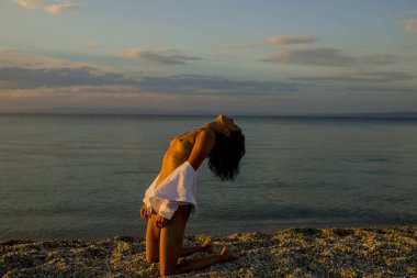 Kadın diz sahilde akşamları durumda. Kız seksi, üstsüz çıplak göğüsler sahil gün batımında, vahşi saçlı. Erotik kavramı. Çekici genç bayan bronzlaşma çıplak, Çıplaklar, son güneş ışınları keyfini çıkarın.