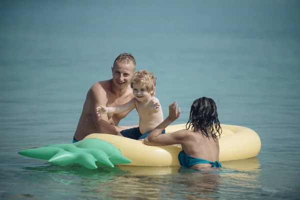 父とマットレスに近い母は、息子と一緒に泳ぐ。家族は、一緒に時間を過ごす楽しんでいます。エアマットレス パイナップル形の海、海、親にかわいい子少年が座っています。家族での休暇の概念. — ストック写真