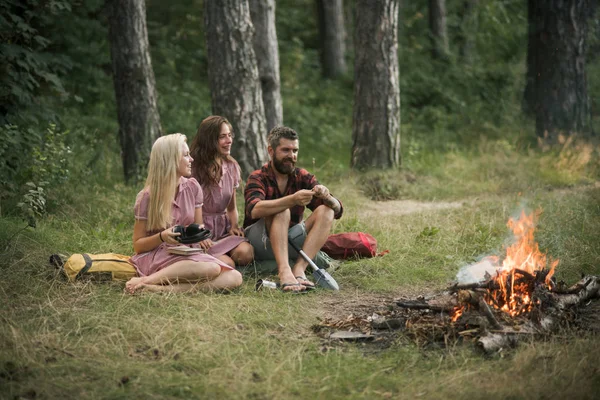 Amigos felizes curtindo piquenique na floresta. Homem barbudo e duas irmãs em vestidos retro sentados ao lado da fogueira e cozinhar comida ao ar livre — Fotografia de Stock