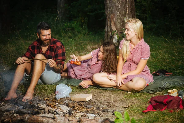 Lächelnder Mann mit stylischem Bart füttert seine brünette Freundin mit über Feuer gekochten Würstchen, Ernährungs- und Diätkonzept. Gruppe fröhlicher Freunde beim Picknick im Wald — Stockfoto