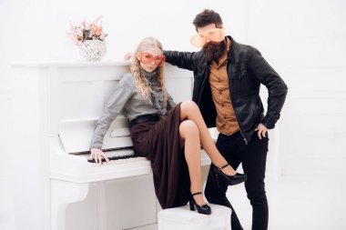 Deri ceket ayakta yanında sakallı adam beyaz piyano üzerinde oturan sarışın kız. İki beyaz studio poz büyük turuncu gözlük içinde