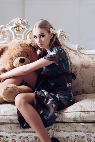 Schöne blonde Mädchen umarmt riesige Teddybär, während sie auf Retro-Sofa sitzen. schlanke Frau im geblümten grauen Kleid in ihrem Zimmer — Stockfoto