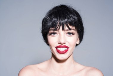 Kadın çekici kırmızı dudaklar ile kameraya benziyor. Siyah peruk Bayan gri arka plan üzerinde olun. Güzel gülümseme kavramı. Kız çıplak omuz ile poz mutlu güler yüz.