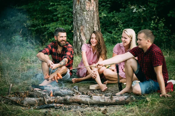 Casais jovens fazendo piquenique na floresta. Homem barbudo e o seu melhor amigo a fritar salsichas em fogo. Pessoas felizes sentadas em torno de fogueira, amizade e conceito de lazer — Fotografia de Stock