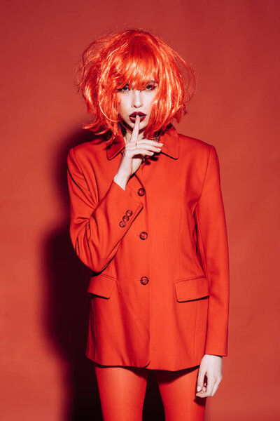 Fashion concept. Lady with dark lips looking at camera with silence gesture. Girl on mysterious face in red formal jacket, red background. Woman with makeup and red wig posing in total red outfit.