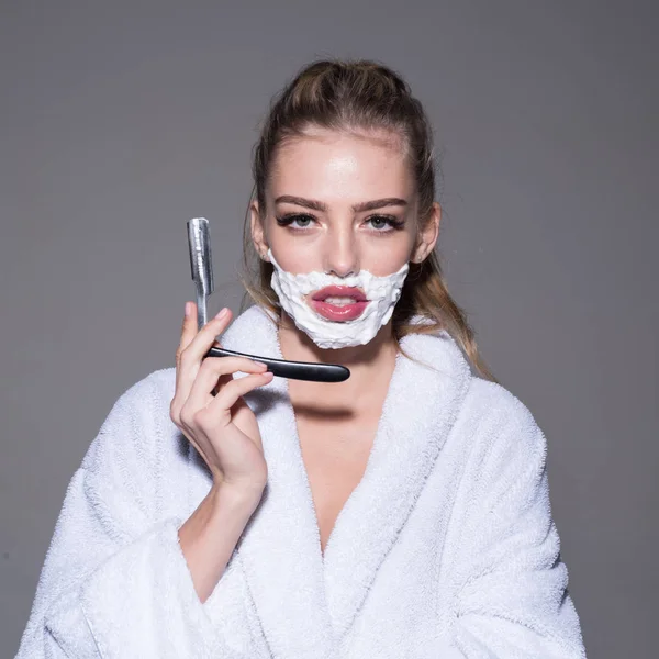 Girl on calm face wears bathrobe, grey background. Woman with face covered with foam holds straight razor in hand. Lady play with sharp blade of straight razor. Barber and shaving concept. — Stock Photo, Image