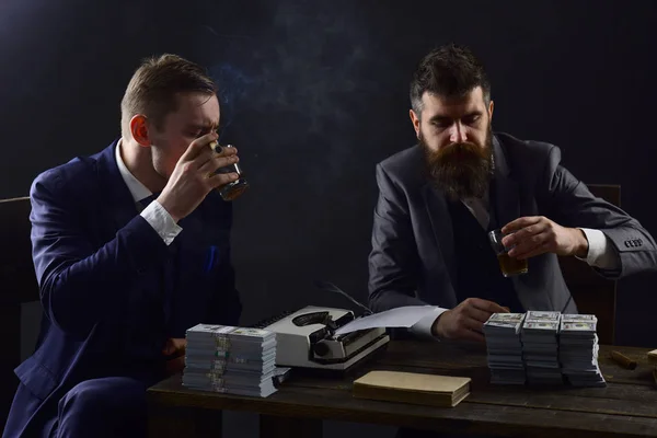 Illegal business concept. Company engaged in illegal business. Men sitting at table with piles of money and typewriter. Businessmen discussing illegal deal while drinking and smoking, dark background.