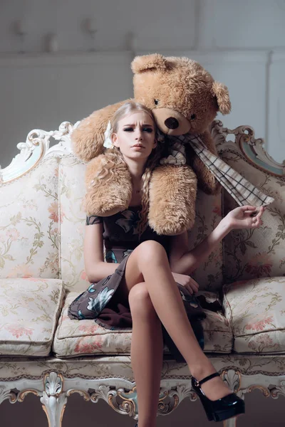 Молодий зіпсований аристократ сидить на старовинному дивані. Блондинка грає з її величезний плюшевий ведмідь — стокове фото