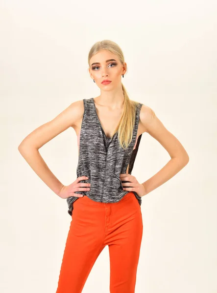 Skinny model with thick eyebrows and neat ponytail wearing melange gray sleeveless blouse and bright orange pants isolated on white background