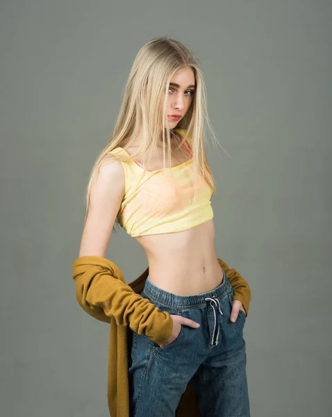 Blond model in yellow top and boyfriend style jeans posing with her hands in pockets isolated on gray background — Stock Photo, Image