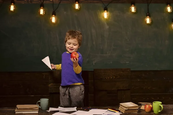 Χαμογελαστό αγόρι ψάχνει στο νόστιμο μήλο στο χέρι του. Ξανθό παιδί κρατώντας χαρτί αεροπλάνο. Ευτυχισμένο παιδί στέκεται πίσω από το τραπέζι — Φωτογραφία Αρχείου