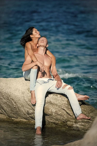 Sexy pareja en topless se sienta en piedra o arrecife en el mar. Familia feliz pasar tiempo juntos, abrazos y bronceado, fondo de la superficie del mar. Concepto de luna de miel. Sensual pareja atractiva desnuda en caras pensativas . — Foto de Stock