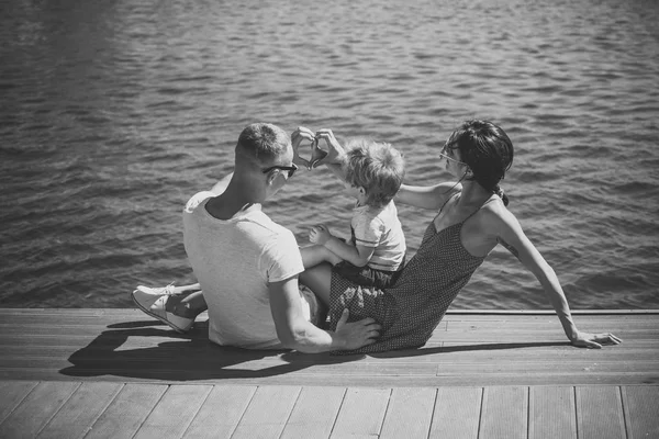 Famiglia felice trascorrere del tempo insieme, sfondo del mare. I genitori con figlio si siedono sul lungomare, vista posteriore. Madre e padre fanno il gesto del cuore o dell'amore con le mani vicino al loro bambino. Concetto vacanza famiglia . — Foto Stock