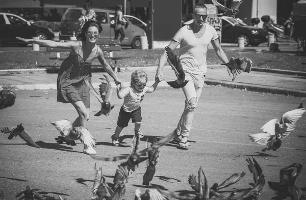 Famiglia felice trascorrere del tempo insieme, sfondo urbano. Genitori con figlio che corre vicino alle colombe, insegue piccioni, ragazzo felice con la faccia sorridente. Madre e padre con il loro bambino. Concetto Family Walk . — Foto Stock