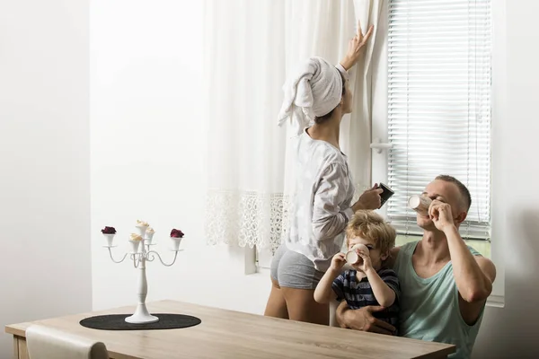 Morning routine concept. Mother and father and child sit at table in morning. Happy family spend time together,white interior and window on background. Family with smiling drinking tea, coffee.