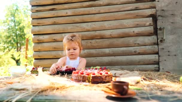 Cute little boy in white pinafore eats fruit cake and cup of milk at table outdoors on wooden. — Stock Video