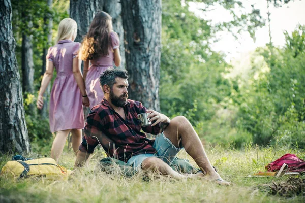 Brodaty mężczyzna picia kawy lub herbaty przy ognisku. Dwie dziewczyny w retro sukienki spaceru na ścieżki w lesie, zawrócić. Koncepcje i relaks — Zdjęcie stockowe
