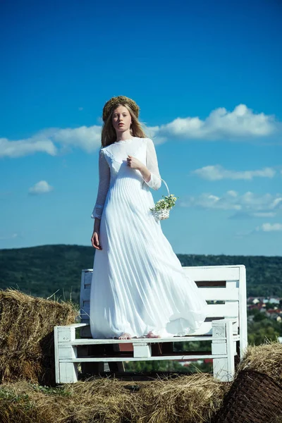 Mulher noiva em vestido de noiva no banco de madeira. Mulher sensual em grinalda no cabelo loiro longo. Albino menina com flores, beleza natural. Modelo de moda no céu azul ensolarado em férias — Fotografia de Stock