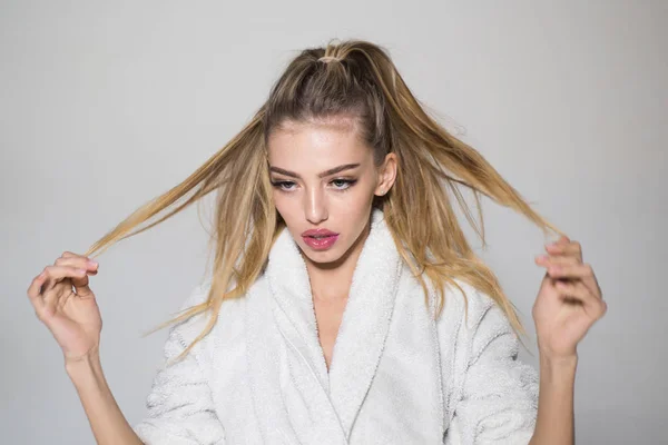 Girl on calm face in bathrobe touches her long hair, grey background. Woman prepares for spa procedures and ties hair in tail. Lady cares about beautiful hair. Spa and wellness concept.