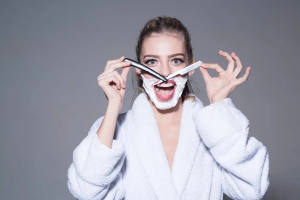 Lady play with sharp blade of straight razor. Woman with face covered with foam holds straight razor in hand. Barber and shaving concept. Girl on smiling face wears bathrobe, grey background. — Stock Photo, Image