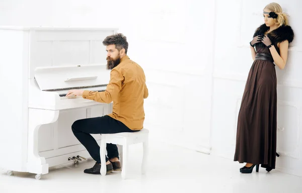 Бородатый мужчина в горчично-желтой рубашке играет на пианино в то время как красивая девушка в платье двадцатых годов и меховой воротник опирается на стену изолированы на белом фоне — стоковое фото
