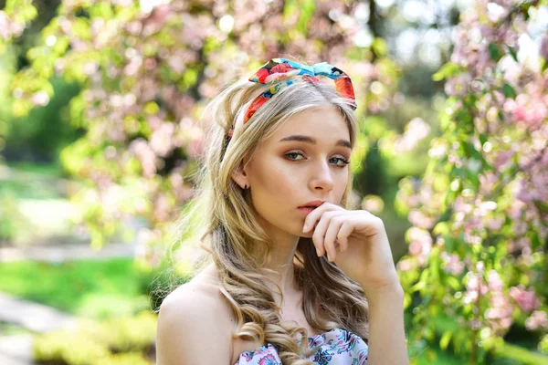 Mädchen auf verträumtem, nachdenklichem Gesicht, zarte Blondine in der Nähe von Sakura-Blumen, Naturhintergrund. Frühjahrsblüher Konzept. Dame spaziert an sonnigem Frühlingstag im Park. junge Frau mit Schal auf dem Kopf genießt die Natur. — Stockfoto