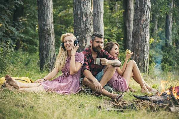 Homme réfléchi avec barbe boire du thé de café tout en regardant les flammes du feu de camp. Fille blonde écoutant de la musique pendant que sa sœur mange des pommes. Diverses perceptions de relaxation parfaite — Photo