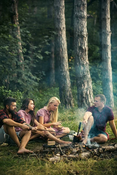 Freunde unterhalten sich am Lagerfeuer. Männer in Holzfällerhemden, die draußen Würstchen backen. Zwei Paare zelten im Wald — Stockfoto