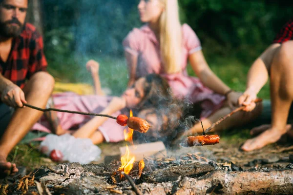 Cuire des saucisses sur des bâtons au-dessus des flammes du feu de camp, camper toute la nuit en été. Groupe d'amis assis par le feu de joie — Photo