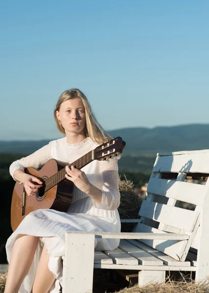 Albino flicka håller akustisk gitarr, stränginstrument. Mode musiker i vit klänning på soliga natur. Sensuell kvinna spela gitarr på träbänk. Kvinna gitarrist utföra musik konsert — Stockfoto