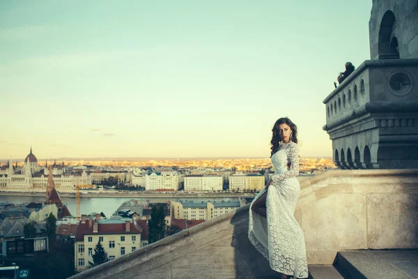 Chica con mirada glamurosa. Mujer en vestido de novia blanco en vista de la ciudad, la moda. Mujer sensual con pelo largo en balcón, belleza. Novia con maquillaje. Modelo de moda estilo y peinado —  Fotos de Stock