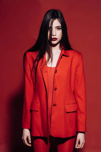 Kvinna med snygg makeup och långt hår poserar i totala röd outfit. Fashion-konceptet. Flicka på mystiska ansikte i röd formella jacka, röd bakgrund. Damen med mörka läppar tittar på kameran. — Stockfoto