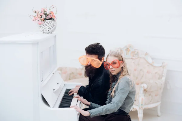 Молода пара у величезних окулярах сидить у білій кімнаті та грає на піаніно. Стильний чоловік з бісеру і його блондинка дівчина сидить на великому піаніно — стокове фото