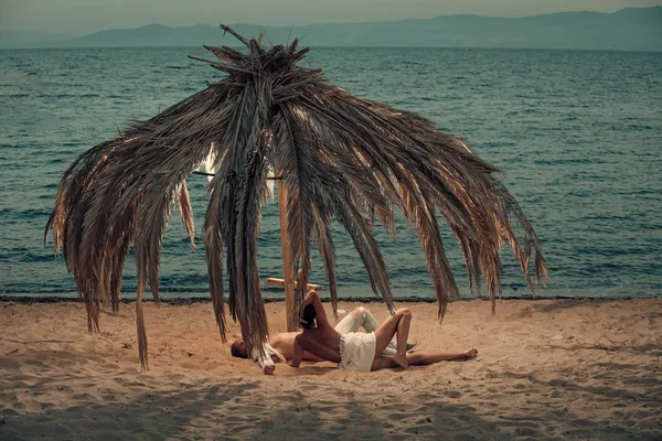 Γυμνόστηθη γυναίκα και άνδρα που βρίσκονται κάτω από την ομπρέλα φτιαγμένο από φύλλα φοίνικα αποξηραμένο. Ζευγάρι σε διακοπές στο ΤΡΟΠΙΚ, μήνα του μέλιτος. Έννοια της αγάπης. Γεμάτος επιθυμία στην παραλία, στη θάλασσα φόντο ερωτευμένο ζευγάρι — Φωτογραφία Αρχείου
