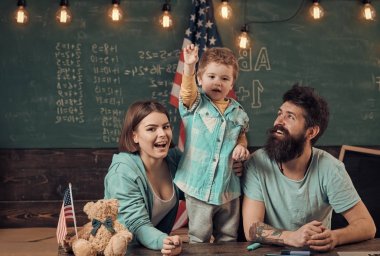 Ebeveynler sınıf ABD bayrağı ile kara tahta arka plan ile evlat. Vatansever eğitim kavramı. Amerikan ailesinin oğlu ve ABD bayrağı ile masa başında oturmak. Anne oğlu Amerikan gelenekleri oynarken öğretim