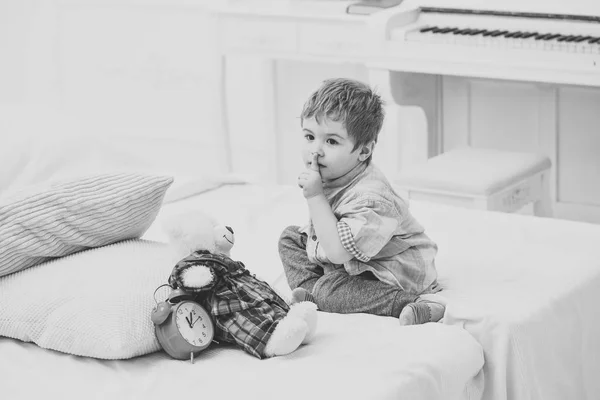 Bambino in camera da letto con gesto di silenzio. Ragazzo messo peluche orso vicino cuscini e sveglia, sfondo interno di lusso. E 'ora di dormire. Ragazzo con la faccia calma mette giocattolo preferito sul letto, tempo di dormire — Foto Stock