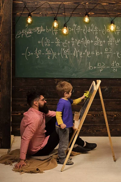 Λίγο μαθηματικά μάθηση ιδιοφυΐα. Δάσκαλος ή ο πατέρας που βοηθούν το παιδί να λύσει εξίσωση στον πίνακα κιμωλίας. Γενειοφόρο σε ροζ πουκάμισο κάθεται στο πάτωμα — Φωτογραφία Αρχείου