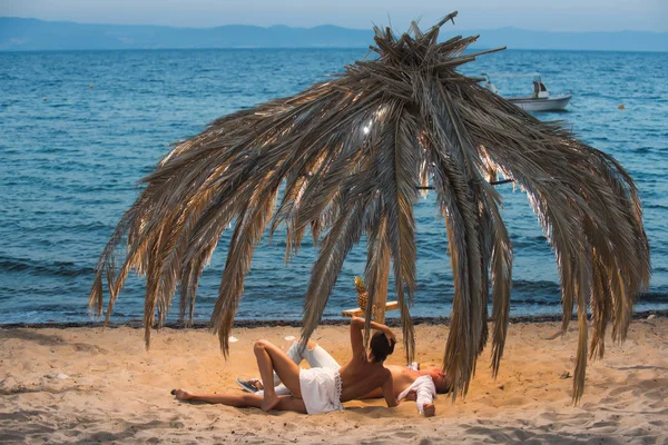 Joven pareja feliz sentada en una arena junto al mar y disfrutando el uno del otro bajo la palmera . — Foto de Stock