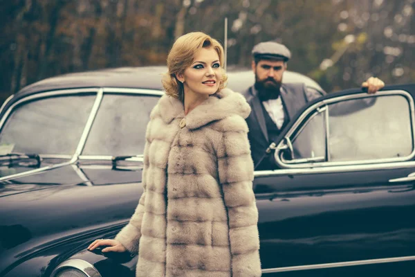 Viajes y viajes de negocios o autoestop. Hombre barbudo y mujer sexy en abrigo de piel. Coche de recogida retro y reparación de automóviles por conductor mecánico. Pareja enamorada en una cita romántica. Escolta de chica por seguridad . — Foto de Stock