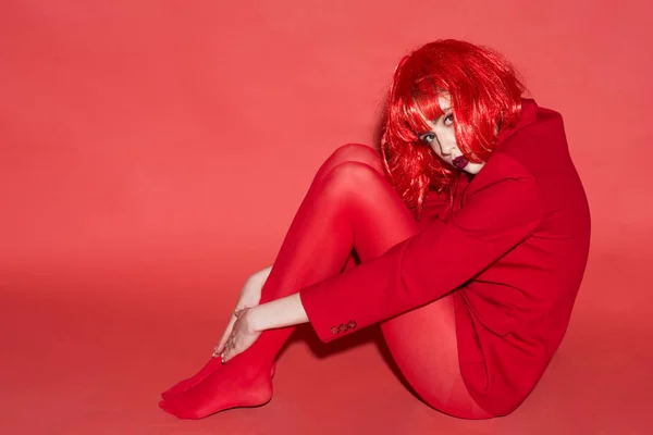 Fashion-konceptet. Damen tittar kameran och sitter på golvet. Kvinna med makeup och röd peruk poserar i totala röd outfit. Flicka på mystiska ansikte i röd formella jacka och tights, röd bakgrund. — Stockfoto