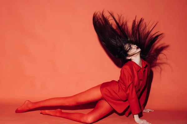 Kvinna med snygg makeup och långt hår poserar i totala röd outfit. Fashion-konceptet. Flicka på mystiska ansikte i röd formella jacka och tights, röd bakgrund. Lady vinka hennes hår medan du sitter på golvet. — Stockfoto