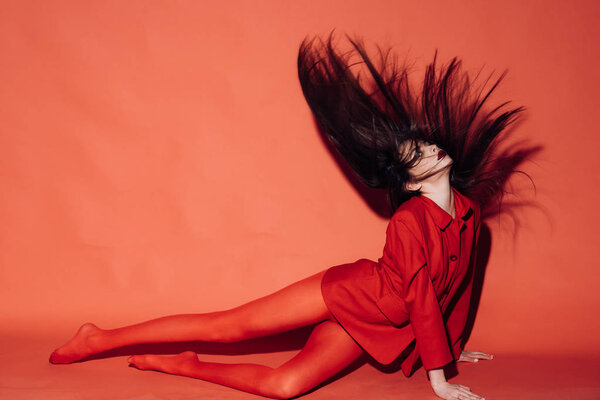 Woman with stylish makeup and long hair posing in total red outfit. Fashion concept. Girl on mysterious face in red formal jacket and tights, red background. Lady waving her hair while sits on floor.