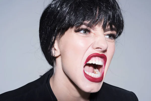 Woman with attractive red lips shouting, close up. Angry boss concept. Girl on scandalous shouting face wears formal jacket. Lady in black wig with make up screaming on grey background.