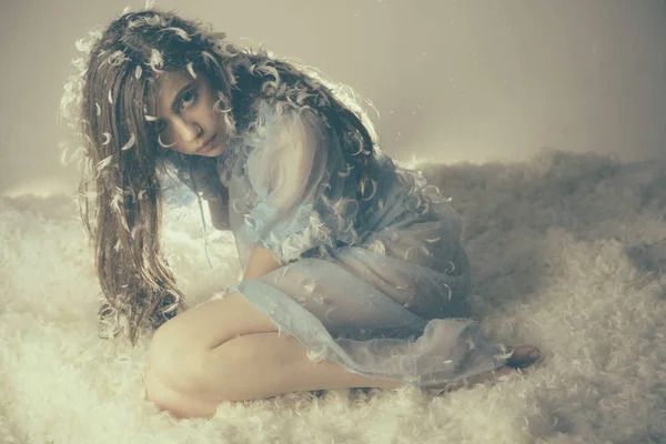 Lady in transparent blue nightie sits on bed while feathers fly around, grey background. Tenderness concept. Girl on calm face sits on bed and play with feather. Woman with long hair in tender pajama. — Stock Photo, Image