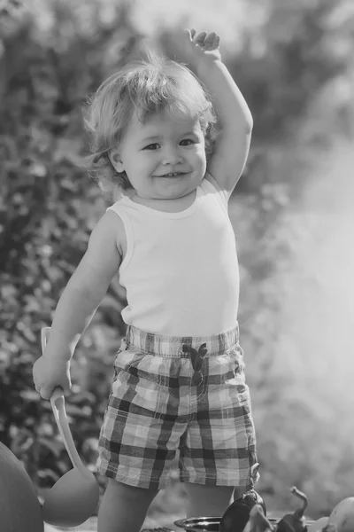 Petit garçon curieux souriant en short d'été à carreaux au pique-nique avec louche debout en plein air sur fond naturel vert journée ensoleillée, image verticale . — Photo