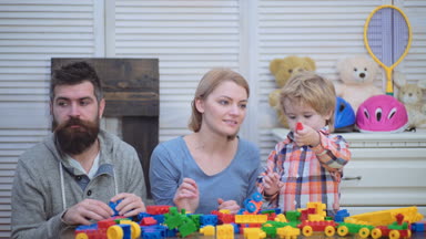 Genç aile inşaat plastik blokları ile oyun oynamak. Aile Oyunları kavramı. Anne ve çocuk mutlu yüzler ile kırmızı tuğla tutun. Baba, anne ve oğlu ışık ahşap arka plan üzerinde oyun odası.
