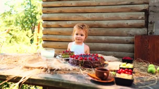 Beyaz önlük içinde şirin küçük çocuk süt bir meyveli kek ve açık havada üzerinde ahşap masada yiyor. — Stok video