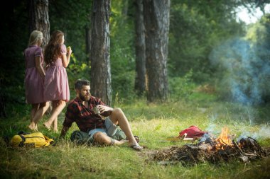 Hippi sakal oturup kamp ateşi ile. Arka plan bulanık kadın ile adam içki çay şenlik ateşi, sakallı. Arkadaşlar ormanda kamp keyfini çıkarın. Yaz tatil kavramı. Kamp ve yürüyüş ve turistik