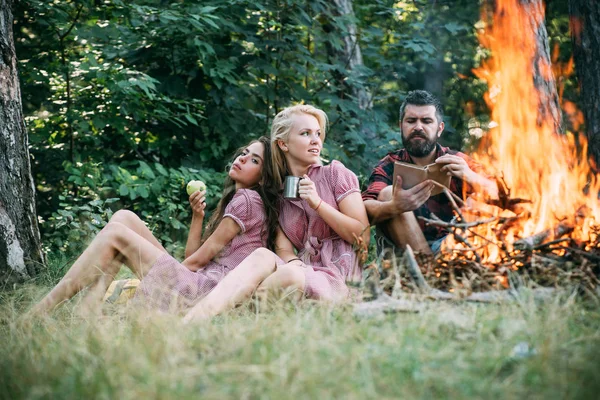 Skupina přátel, kempování v lese. Guy čtení kniha ohněm. Krásná blonďatá dívka pití čaje z hrnek, zatímco její přítel jí zelené jablko — Stock fotografie